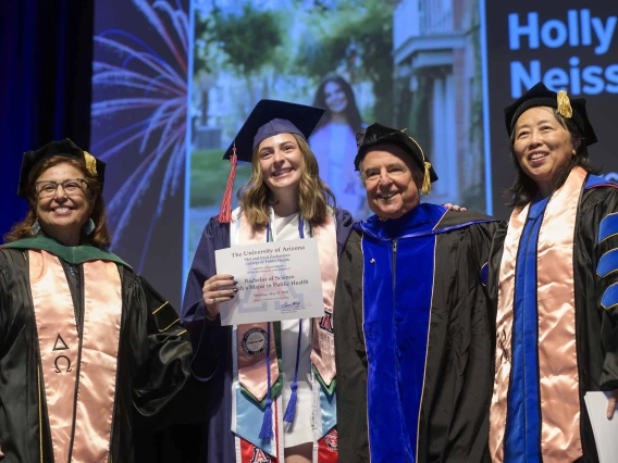 A young woman holds up a certificate and smiles while standing on a stage with several professors. All are wearing graduation regalia. 