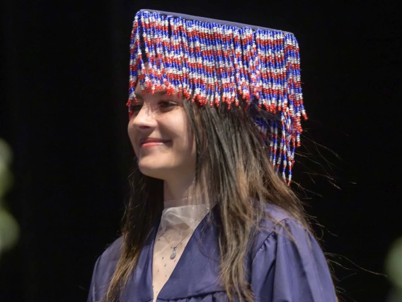 A young woman wearing a graduation gown and with beads hanging from her mortar board walks across a stage, smiling. 