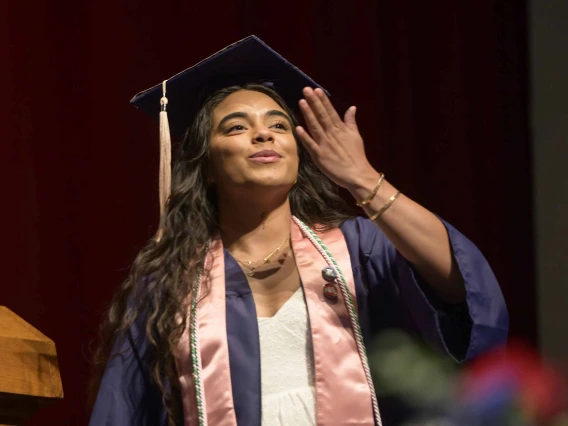 A young woman dressed in a graduation cap and gown blows a kiss as she walks across a stage during a graduation ceremony. 