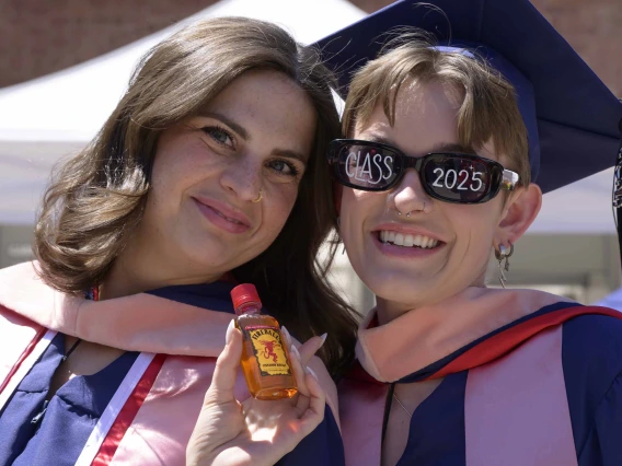 Two young women wearing graduation regalia smile outside after their graduation ceremony. One is wearing sunglasses that read “Class 2025” on the lenses. 