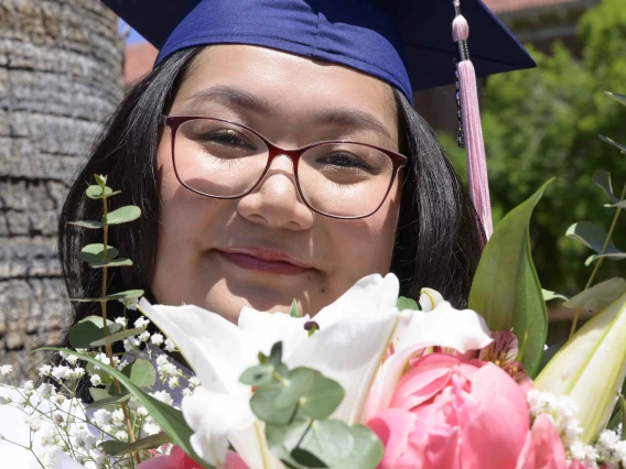 A young woman wearing a graduation cap smiles as she looks over a bouquet of flowers. 