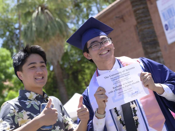 A young man wearing a graduation cap and gown smiles as he holds up his diploma while his friend looks on. 