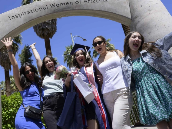 Five young women stand together, smiling and cheering, under a cement arch that reads “Inspirational Women of Arizona” outside. One of the women is wearing a graduation cap and gown. 