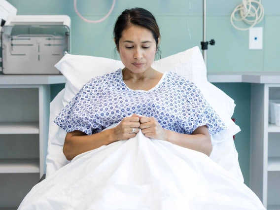 Woman wearing hospital gown laying on gurney with pensive look, clutching blanket with both hands