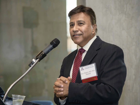 A man in a dark suit speaks into a microphone at a lectern; his hands are clasped in front of him.