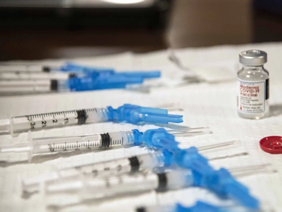 A table covered by a while cloth holding a vial with label “Moderna COVID-19 vaccine” and several syringes with needles ready for vaccination injections