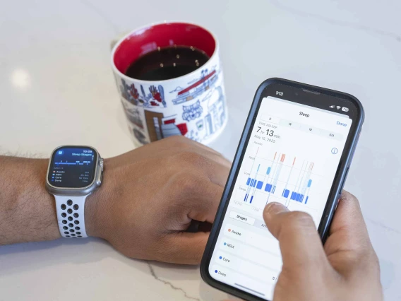 A person’s hands resting on a table, with an Apple Watch on the left wrist and right hand holding an iPhone displaying sleep date. A University of Arizona mug of coffee sits on the table 