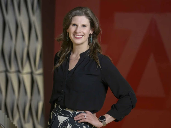 Portrait of professor Rina Fox smiling as she stands in front of a red wall with a large “A” on it. 