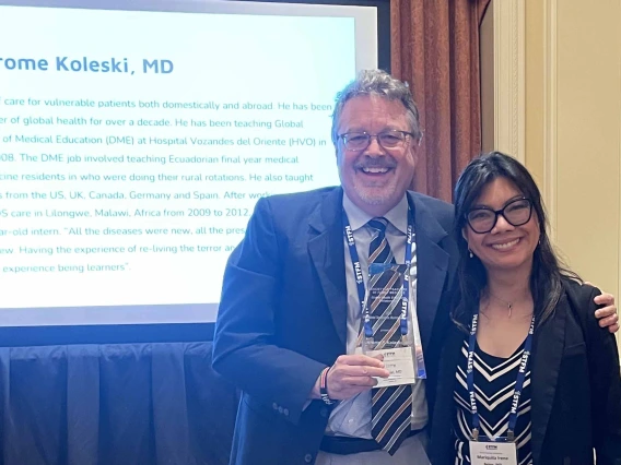 Jerome Koleski, MD, smiles while holding a crystal award as he stands next to a woman in a conference room.