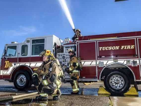 Two firefighters in the foreground and one spraying water from the top of a Tucson Fire firetruck