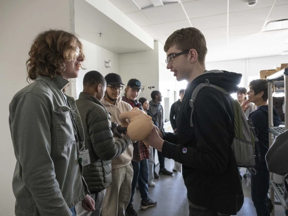 A line of high school students pass a baby doll to each other as they stand in a large room. 