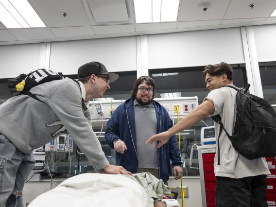 Two high school students laugh as they perform CPR on a manikin while an instructor looks on in a medical training room. 