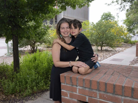 A young boy kneels on a raised surface and reaches out to hug his mother