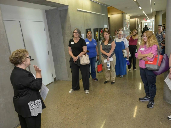 A woman with her back to the camera, one hand raised, speaks to a group of educators gathered outside an elevator at the Phoenix Bioscience Core. 