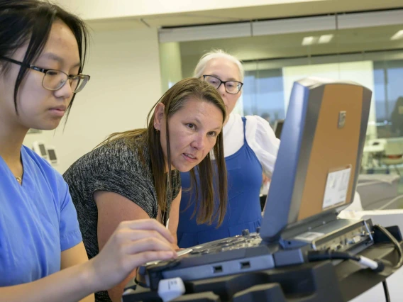 A teacher performing an ultrasound simulation looks at the image, while a fellow teacher watches and a medical student runs the monitor. 