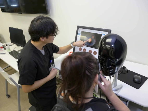 A person with his back to the camera points at a computer screen displaying a virtual eye image while another person peers into an ophthalmology simulator