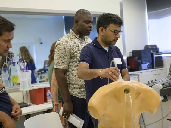 Three educators gather around an ultrasound station where one holds a wand over a manikin, viewing a monitor displaying simulated health conditions, while the others observe. 