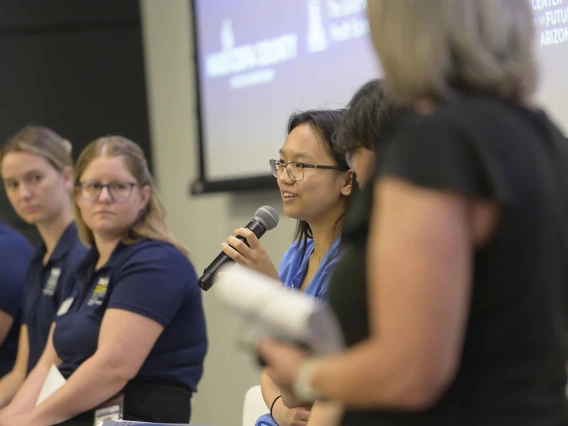 A person holds a microphone, speaking in a conference room, with four people seated on other side of her as part of a panel discussion.