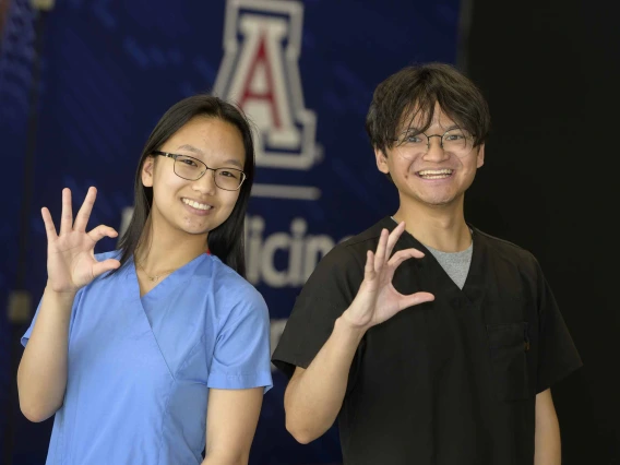 Two people stand side by side, smiling and holding up University of Arizona Wildcat signs with their right hands.