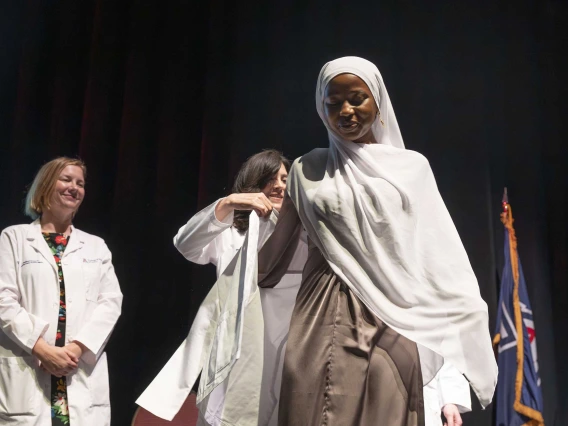 A medical student wearing a hijab is assisted in putting on a medical white coat by a professor in a medical white coat. 