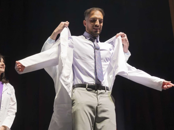 A medical student puts on his white coat with the help of a professor who is standing behind him.