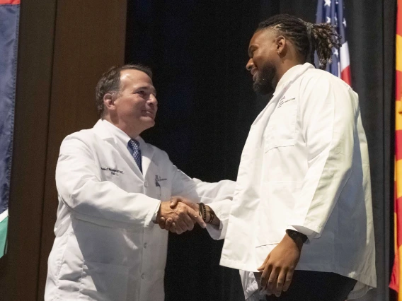Fred E. Wondisford, MD, MS, MBA, shakes hands with a new medical student onstage. Both are smiling and wearing medical white coats. 