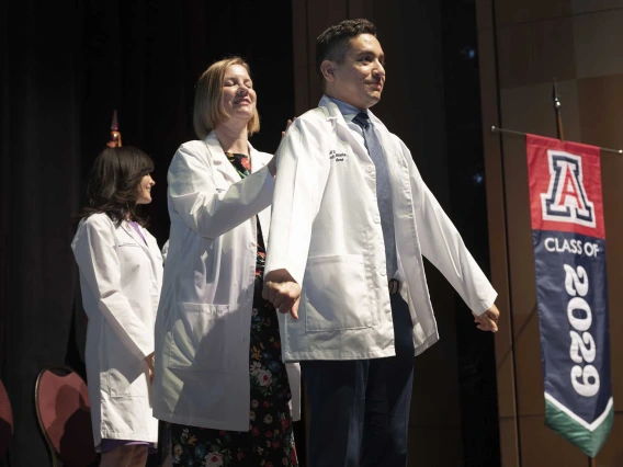 A medical student puts on his white coat with the help of a professor who is standing behind him.