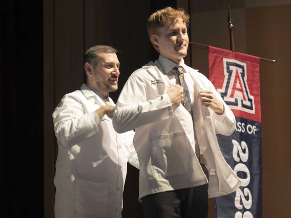 A medical student puts on his white coat with the help of a professor who is standing behind him.