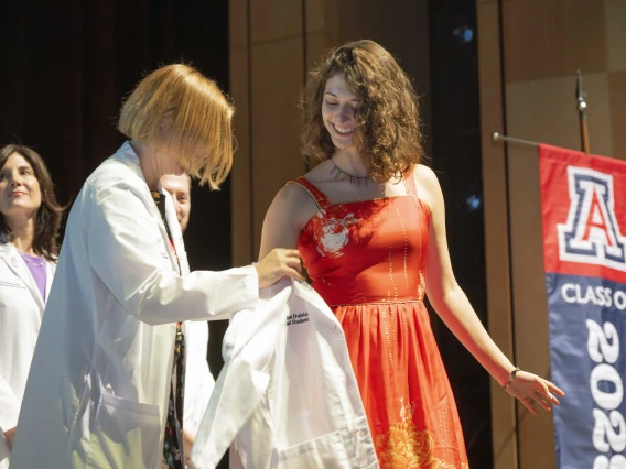 A professor in a medical white coat helps a new medical student put on her white coat during a ceremony. 