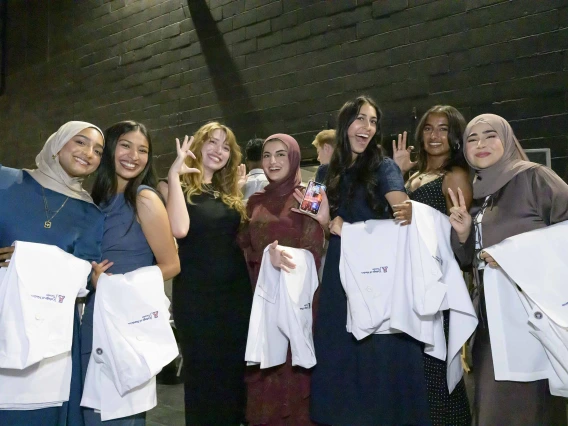 Seven female medical students stand together, smiling, while holding medical white coats over their arms. 
