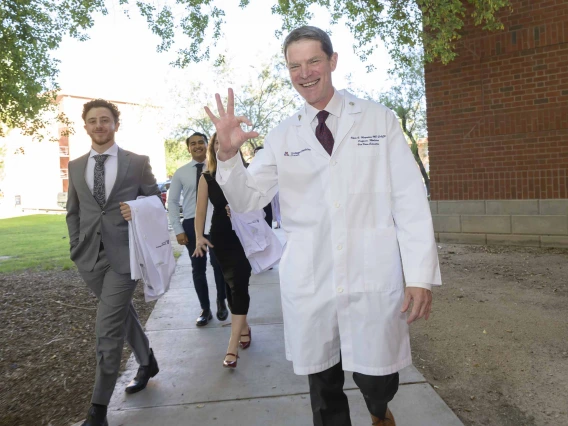 A smiling Professor Kevin F. Moynahan, MD, dressed in a medical white coat, leads a group of first-year medical students down a sidewalk outside a brick building. 