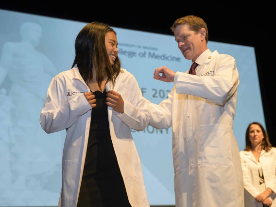 A first-year medical student wearing a medical white coat looks back and smiles at her mentor, a doctor who is also wearing a medical white coat, while standing onstage.  