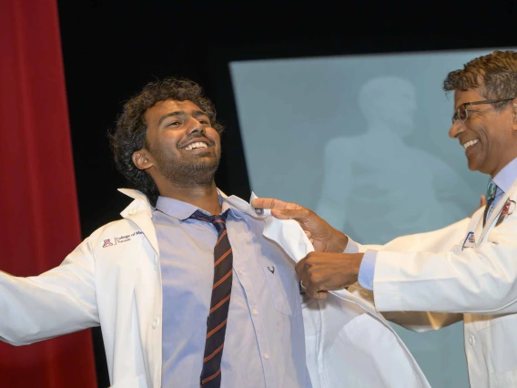 A new medical student laughs as a smiling professor helps him put on his white coat. 
