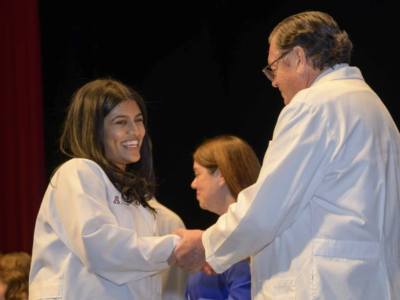 A first-year medical student, smiling and wearing a white medical coat, shakes hands with the dean of the college, who is also wearing a white coat. 