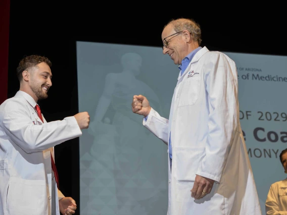 A medical student and his professor, both wearing white coats, smile as they bump fists onstage. 