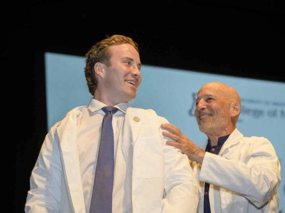 A first-year medical student wearing a medical white coat looks back and smiles at his mentor, a doctor who is also wearing a medical white coat, while standing onstage.  
