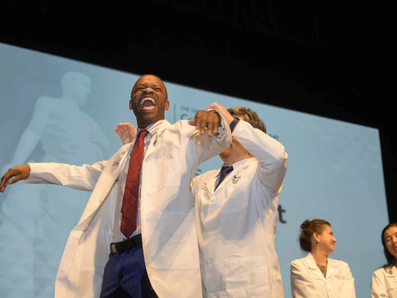 A first-year medical student holds his arms out and laughs as a professor standing behind him helps him put on a medical white coat. 