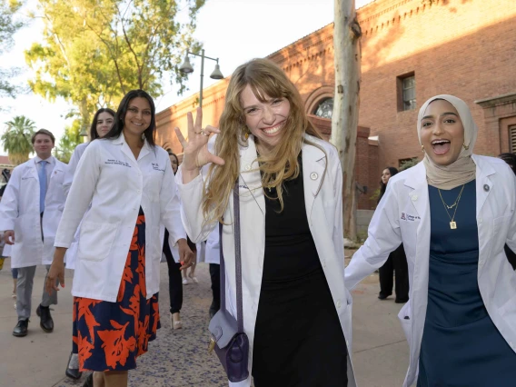 Several first-year medical students wearing white coats smile and celebrate as they walk outside Centennial Hall after their white coat ceremony. 