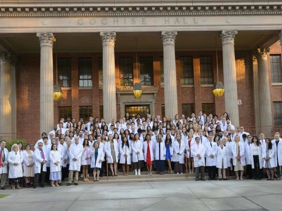 The 120 members of the College of Medicine – Tucson Class of 2029, along with faculty mentors, stand together outside a building with large columns and stairs. All are wearing medical white coats. 