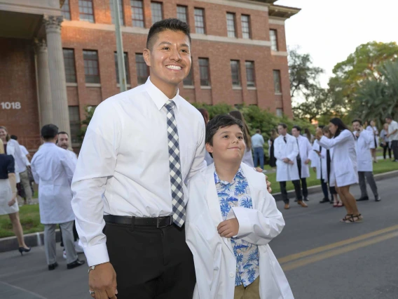 A first-year medical student wearing a shirt and tie stands with his arm around his younger cousin, who is wearing his medical white coat. The coat is very large on the younger cousin. 