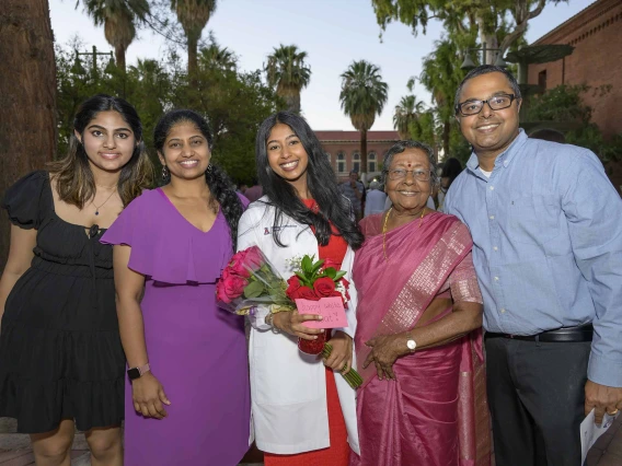 A first-year medical student wearing a white medical coat smiles and holds flowers while standing with smiling family members outside. 