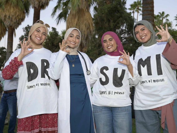Four smiling young women stand together and flash the U of A Wildcat hand sign. One is wearing a medical white coat and the other three are wearing white T-shirts with large black lettering spelling out “Dr. Sumi,” with two large letters on each shirt. 