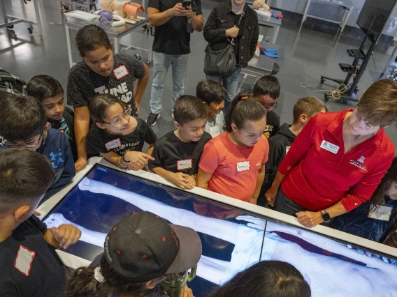 About a dozen grade-school students stand around a table with a digital image of a body while a tour guide demonstrates how it works. 
