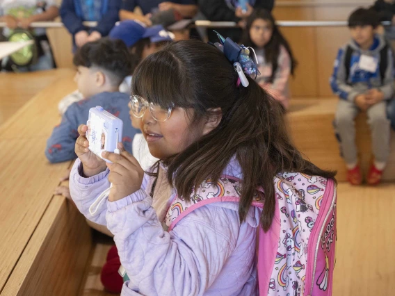 A young girl stands in front of other students while holding up a camera to take a photo. 