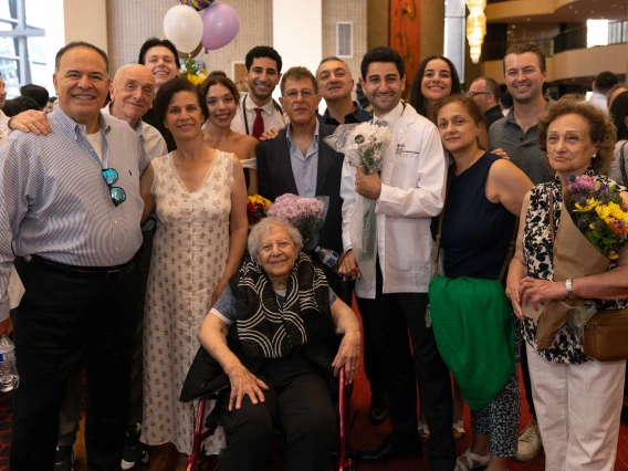 A large family stands around two medical student wearing a white coats in a crowded lobby. 
