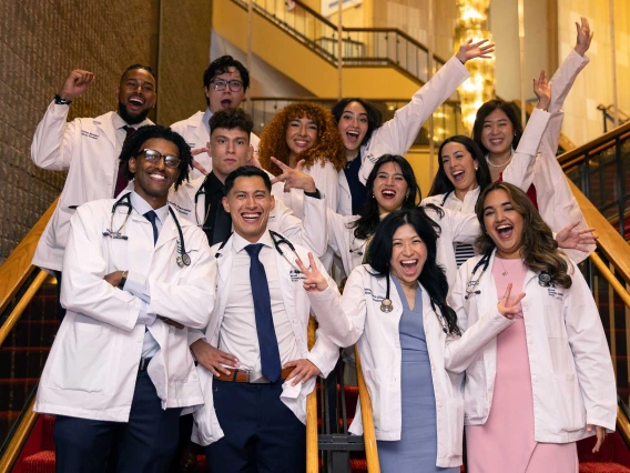 About a dozen students wearing medical white coats stand on the stairs in a lobby, smiling and cheering. 