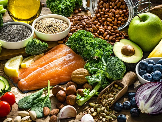 Healthy foods on a table: salmon, kale, spinach, avocado, almonds, blueberries, onion, chia seeds, cucumber, tomato, apple, pistachios, garlic, carafe of olive oil, lemon, broccoli
