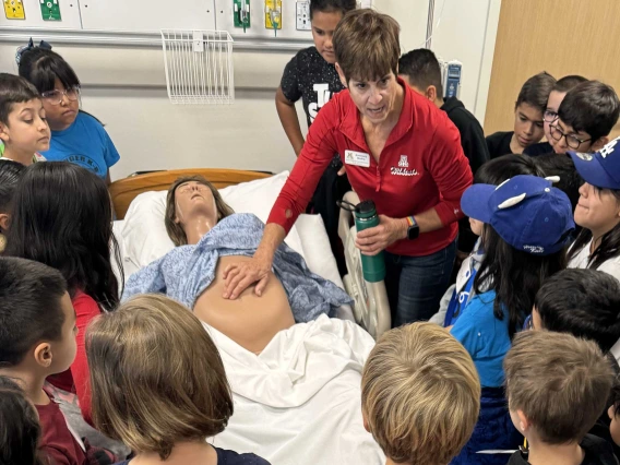 More than 20 elementary school children stand around a pregnant manikin lying on a hospital bed as an adult pushes on the manikin’s abdomen. 