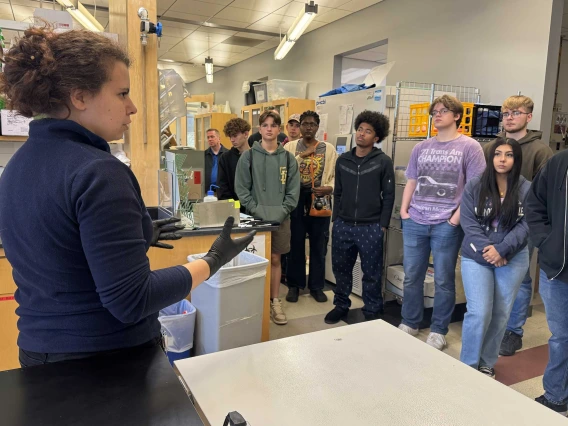 A woman speaks to a group of high school students in a science lab.  
