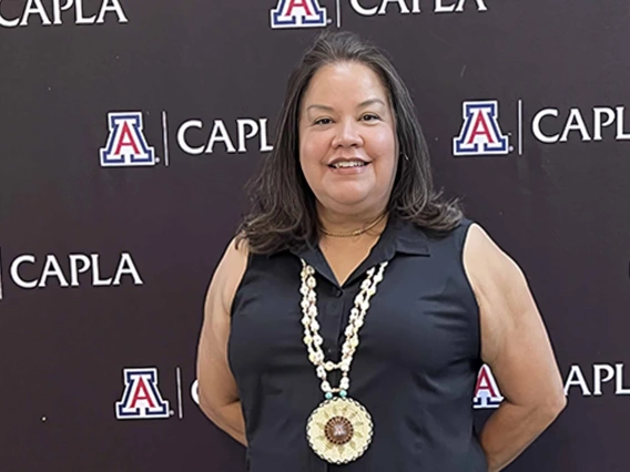 Portrait of Christina C. Bell Andrews, JD, MPH, MBA, MA, with a medal around her neck in front of a background with the University of Arizona College of Architecture, Planning and Landscape Architecture logo.